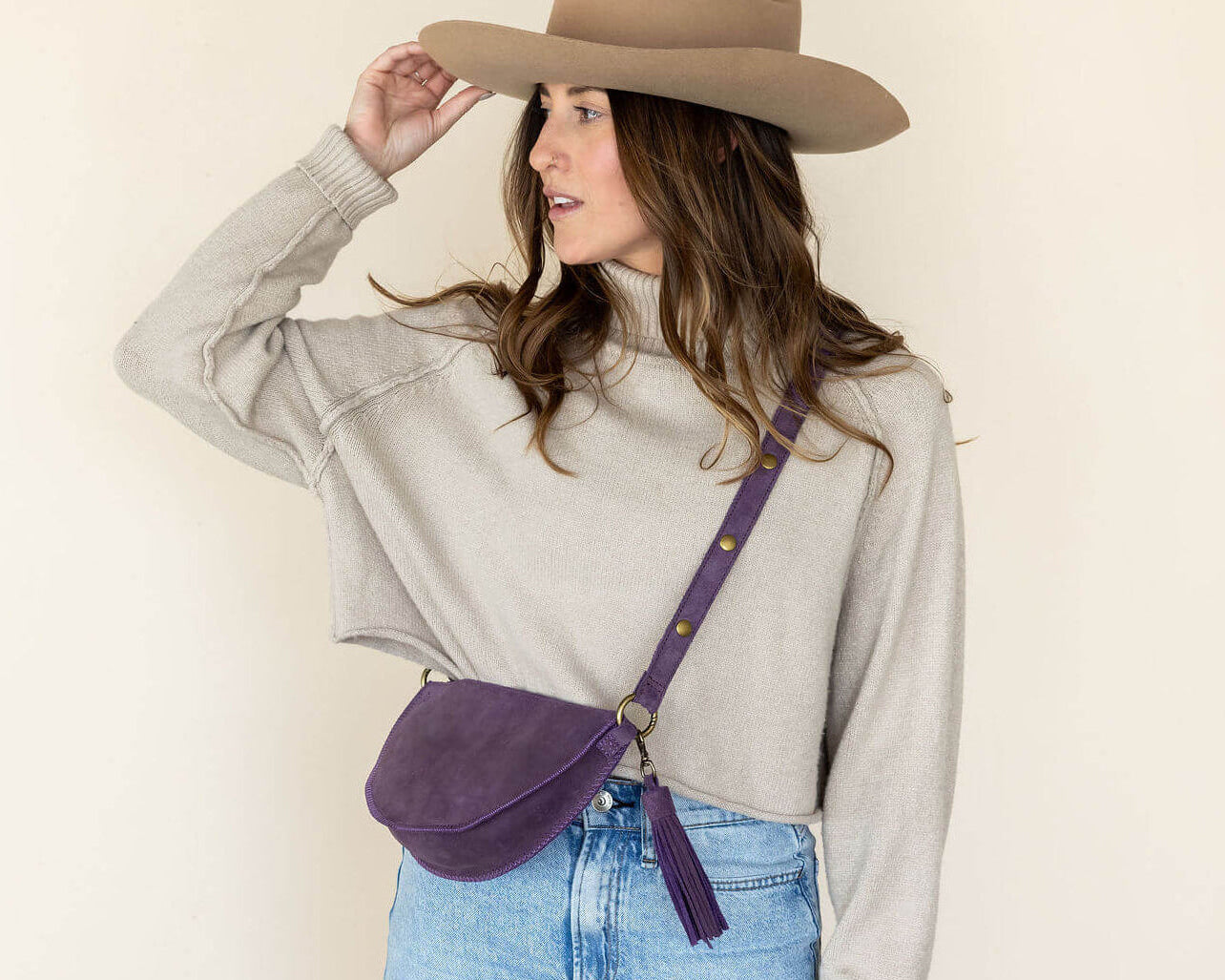 Woman wearing a beige hat and light gray shirt with a purple crossbody bag on a plain background