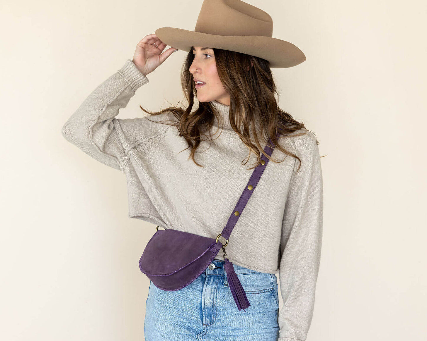 Woman wearing a beige hat and light gray shirt with a purple crossbody bag on a plain background