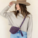 Woman wearing a beige hat and light gray shirt with a purple crossbody bag on a plain background