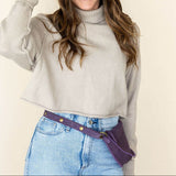 Woman wearing a beige sweater, blue jeans, and a brown hat against a plain background