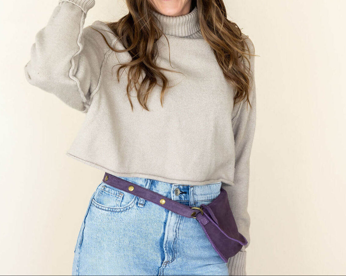Woman wearing a beige sweater, blue jeans, and a brown hat against a plain background