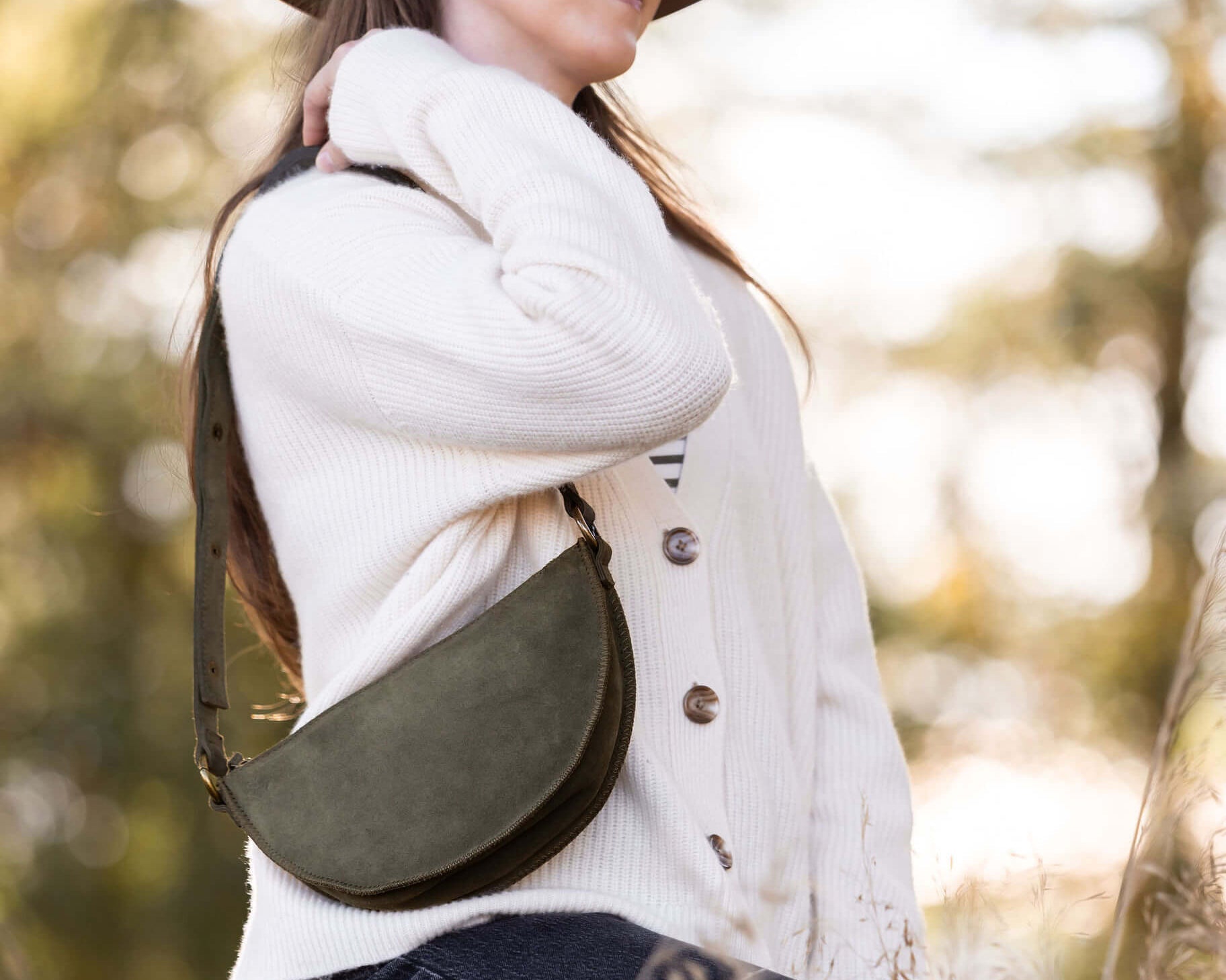 Woman wearing a white sweater and brown hat with a green leather bag over her shoulder, standing in a blurred natural setting.