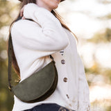 Woman wearing a white sweater and brown hat with a green leather bag over her shoulder, standing in a blurred natural setting.