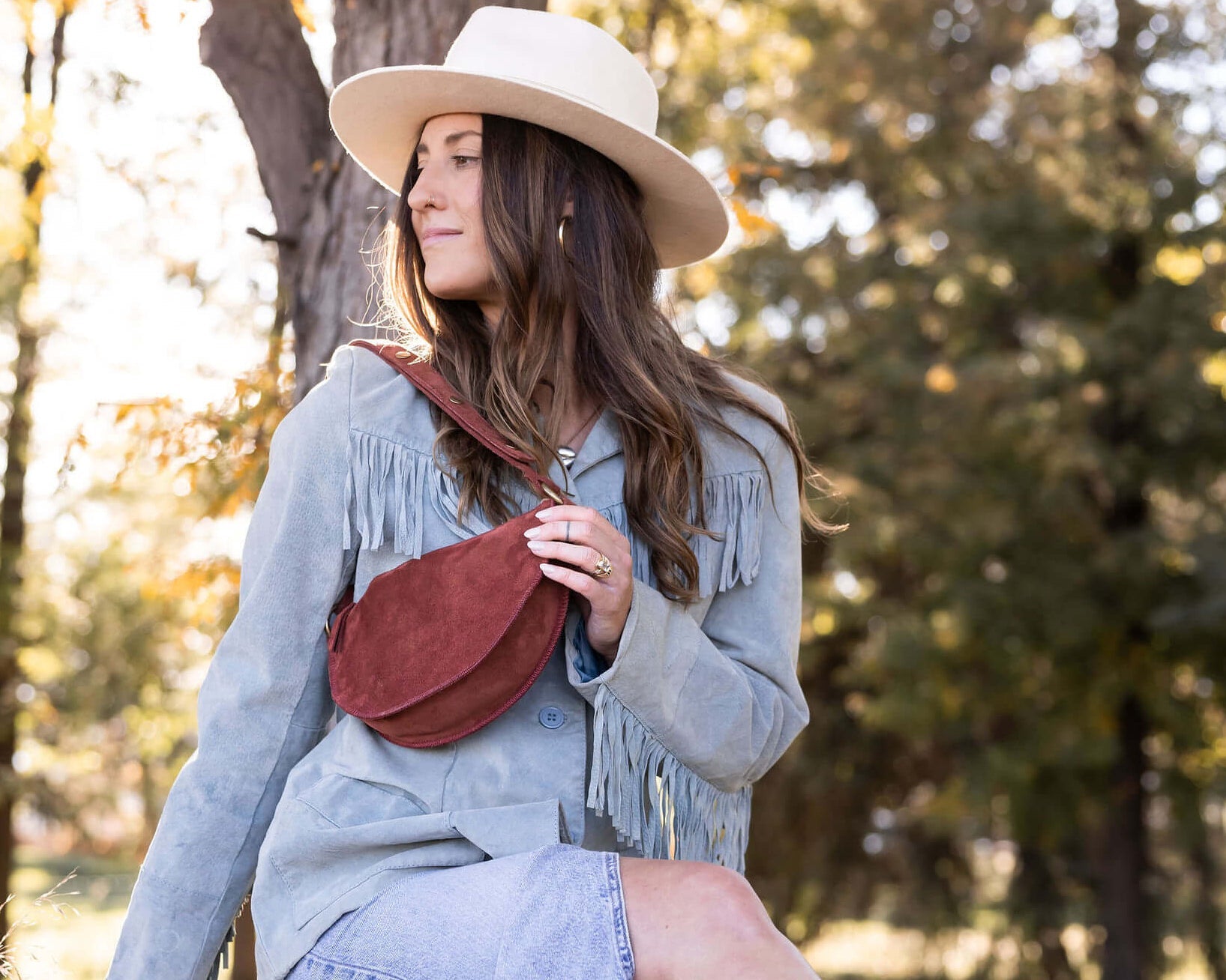 Woman sitting on a wooden fence wearing a light blue coat, red boots, and a beige hat in a natural setting.