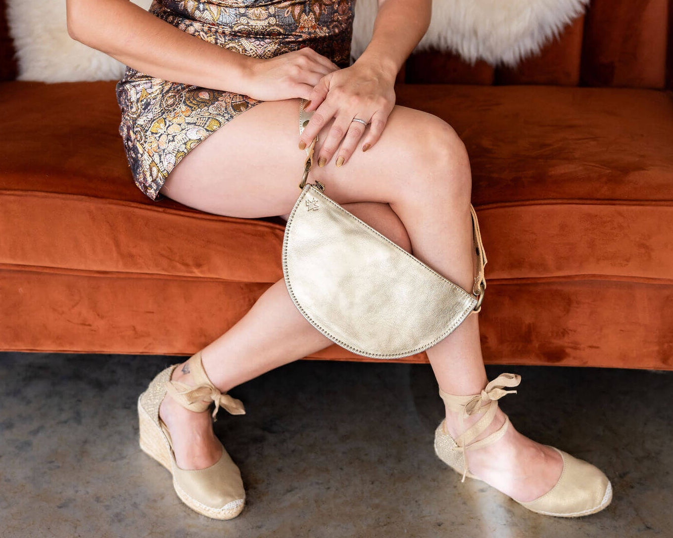 Person sitting on a brown couch holding a gold handbag, wearing a patterned dress and beige shoes.