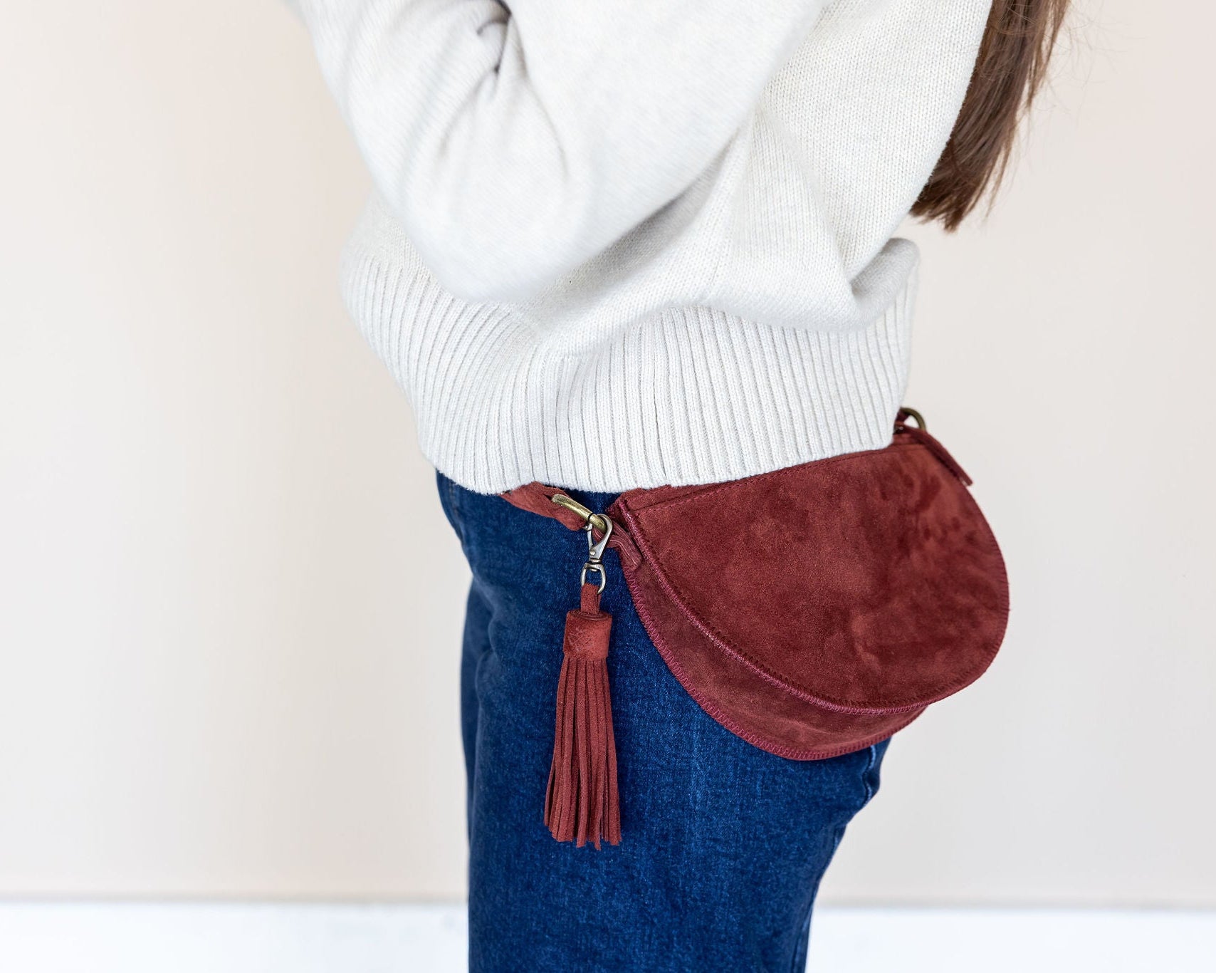 Person wearing a white sweater and blue jeans with a red suede bag on a beige background