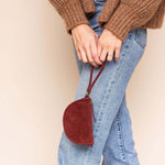 Person wearing a brown sweater, blue jeans, and brown boots holding a red handbag against a beige wall.