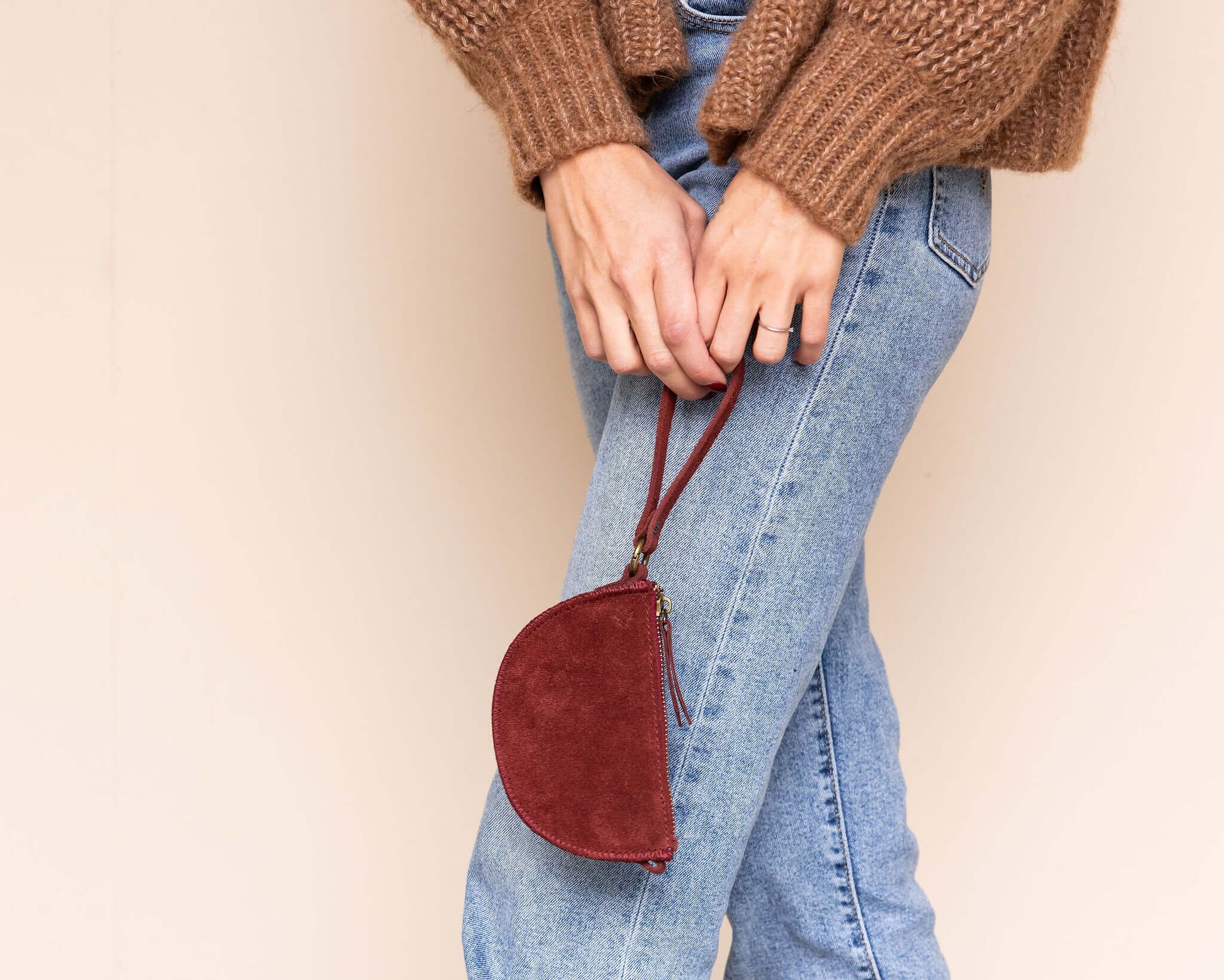 Person wearing a brown sweater, blue jeans, and brown boots holding a red handbag against a beige wall.