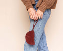 Person wearing a brown sweater, blue jeans, and brown boots holding a red handbag against a beige wall.