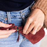 Person adjusting a red leather strap on a bag, wearing a brown coat and blue jeans.