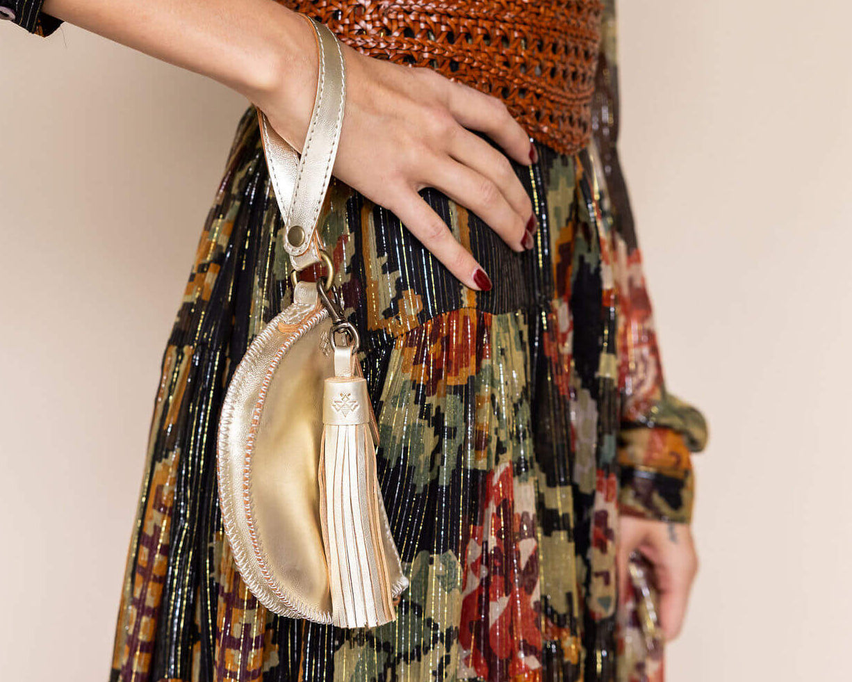 Person wearing a patterned dress holding a gold handbag with a tassel on a plain background