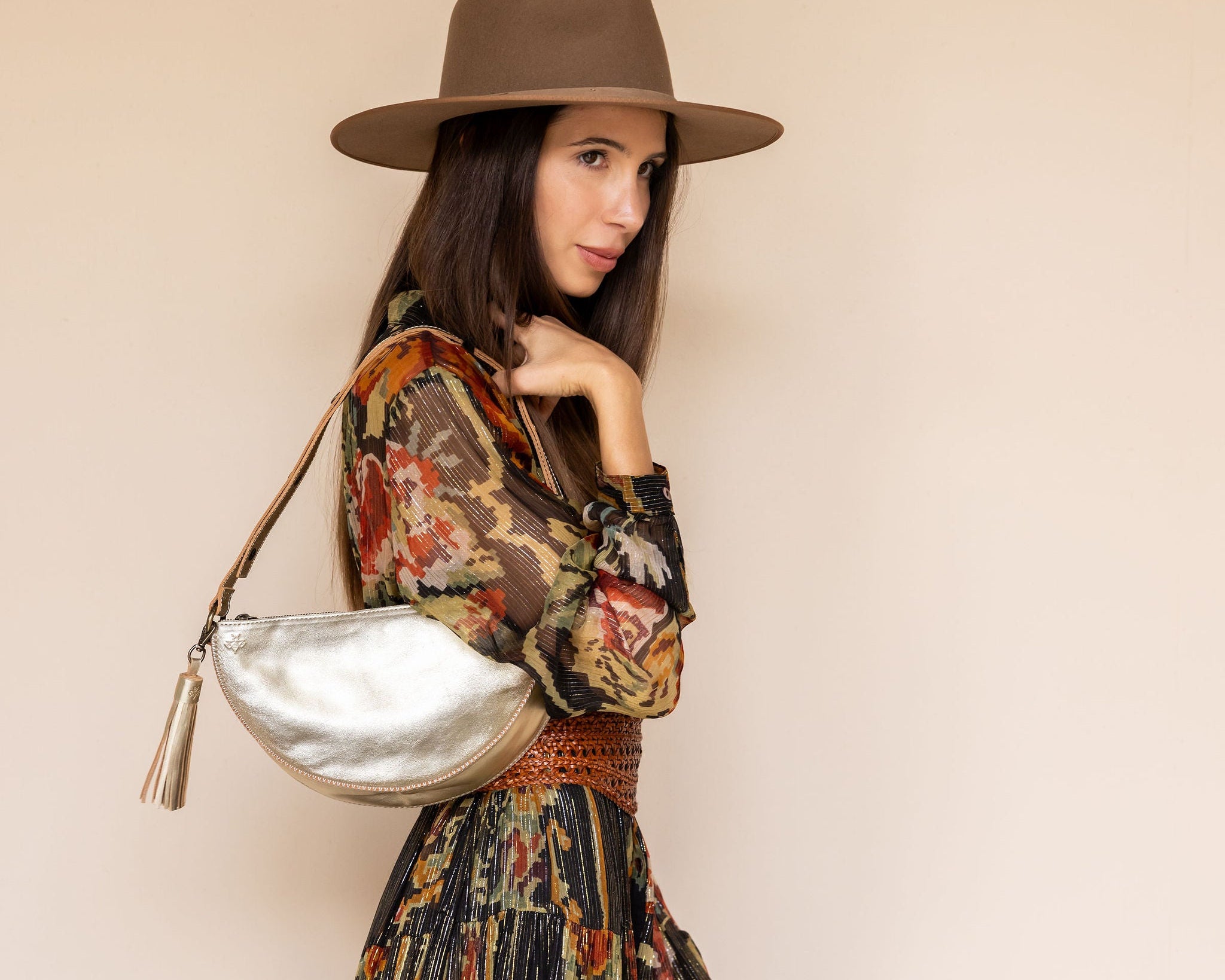 Woman wearing a patterned dress, brown hat, and holding a silver handbag against a beige background