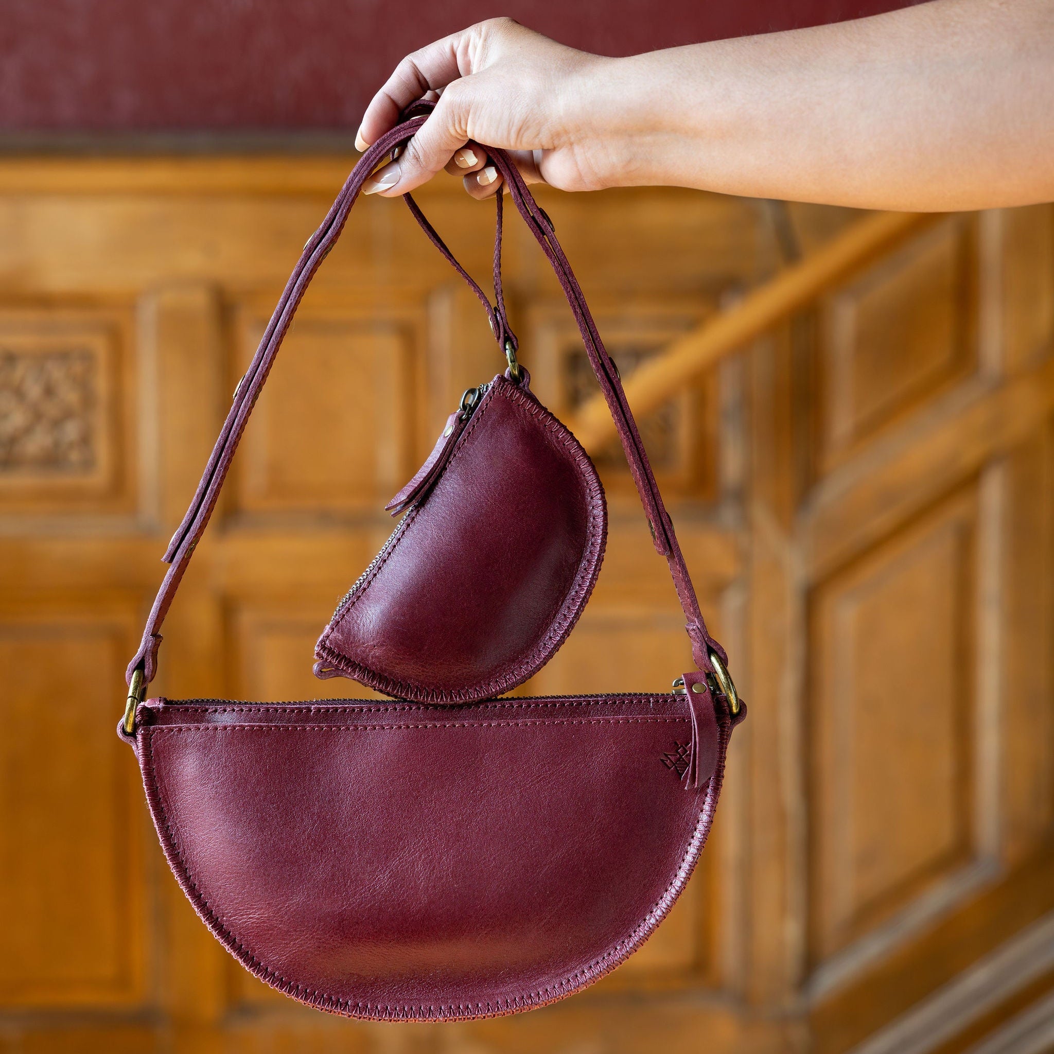 Hand holding a burgundy leather handbag against a wooden wall.