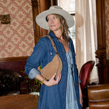Woman in a blue dress and hat standing in an ornate room with patterned wallpaper and leather furniture.