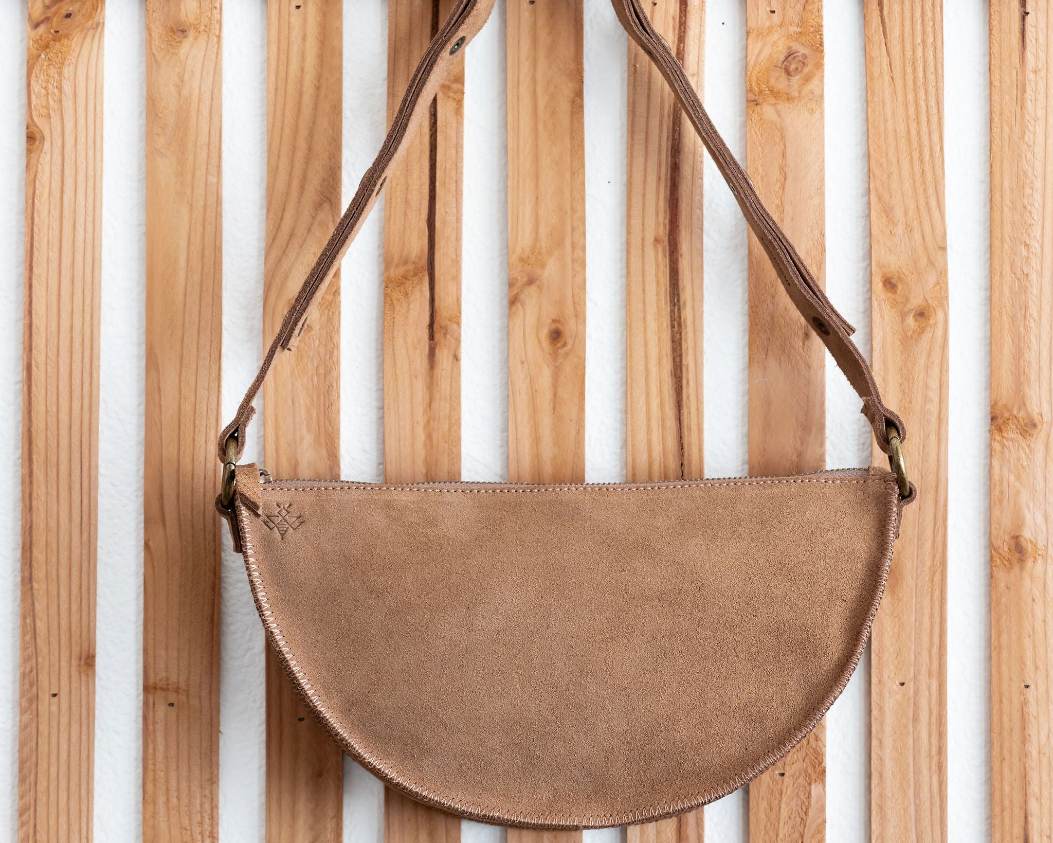 Brown suede handbag with leather strap hanging on a wooden slatted wall.