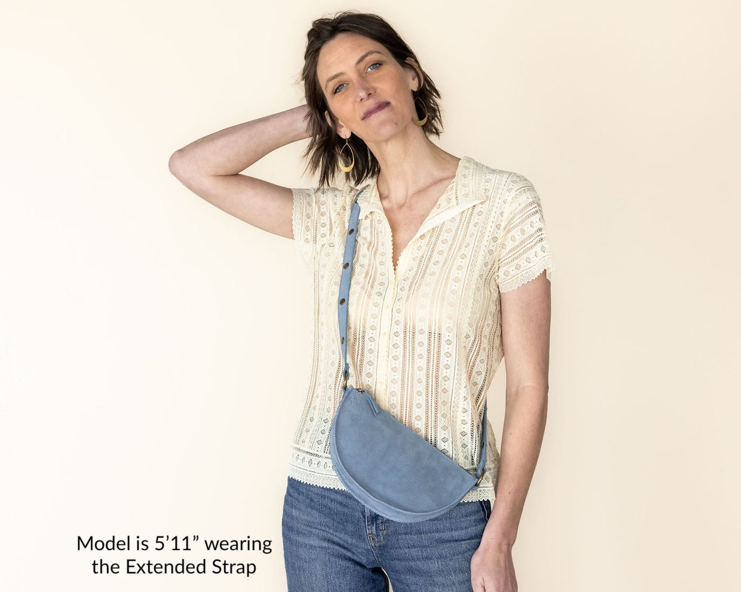 Woman wearing a beige blouse and blue jeans with an extended strap over her shoulder on a white background