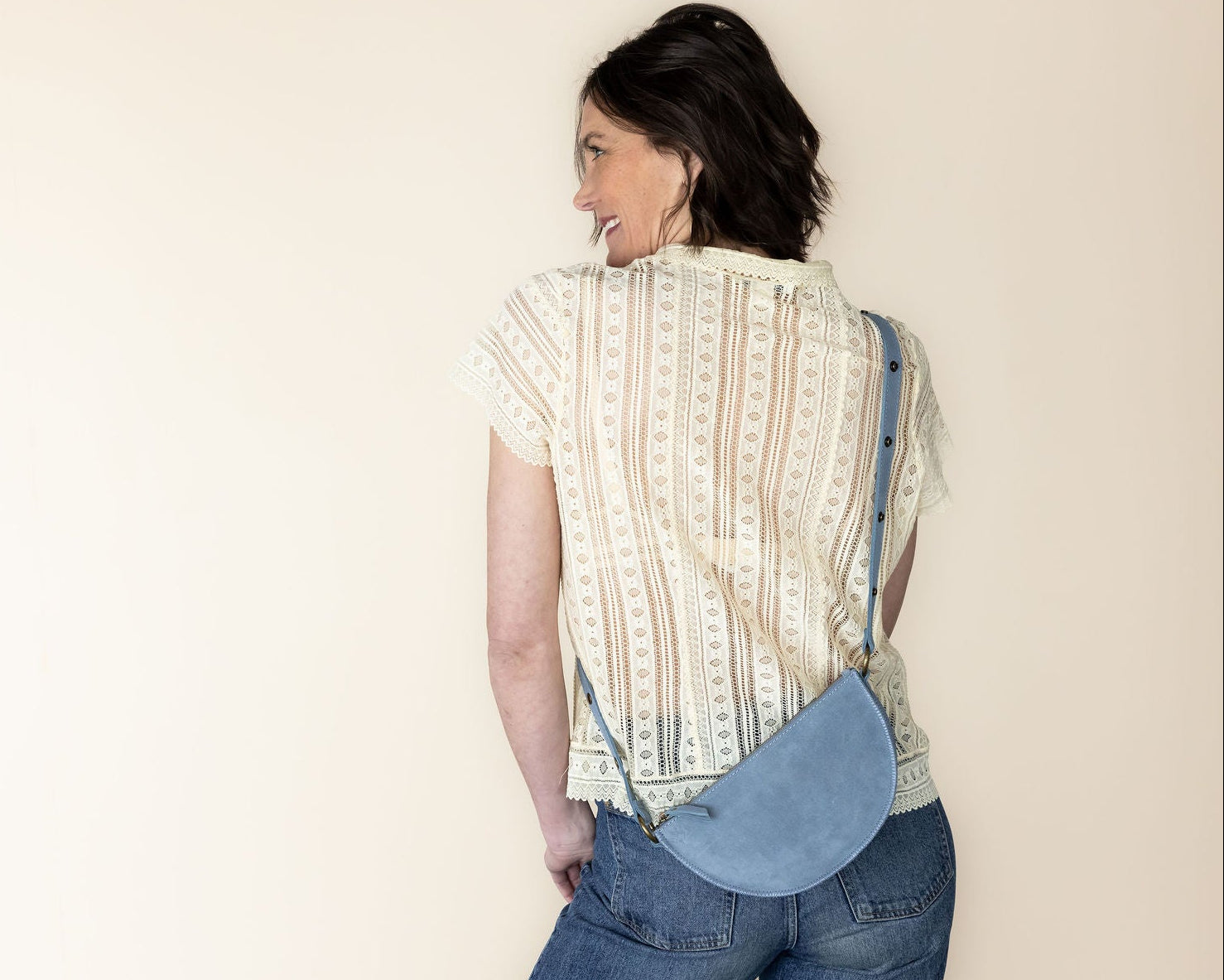 Woman wearing a patterned shirt and jeans with a blue bag, standing against a plain background