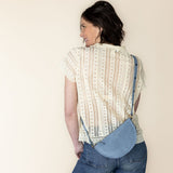 Woman wearing a patterned shirt and jeans with a blue bag, standing against a plain background
