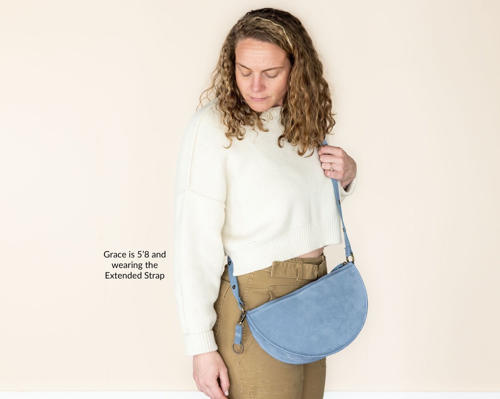 Woman holding a blue crossbody bag with an extended strap against a beige background