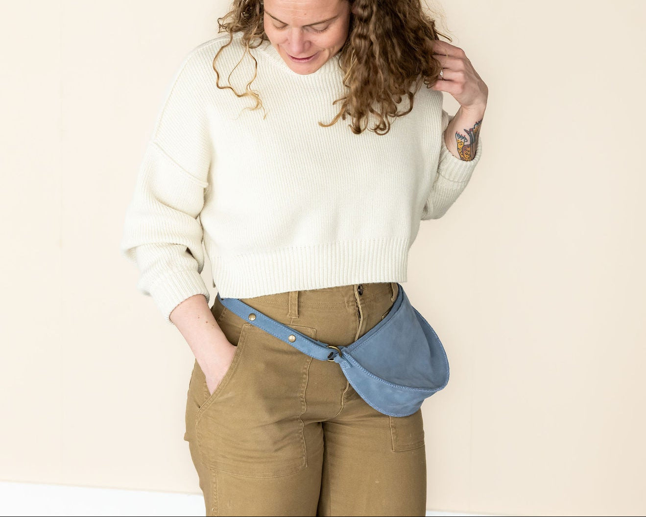 Woman wearing a white sweater and brown pants with a blue fanny pack against a beige background