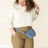Woman wearing a white sweater and brown pants with a blue fanny pack against a beige background
