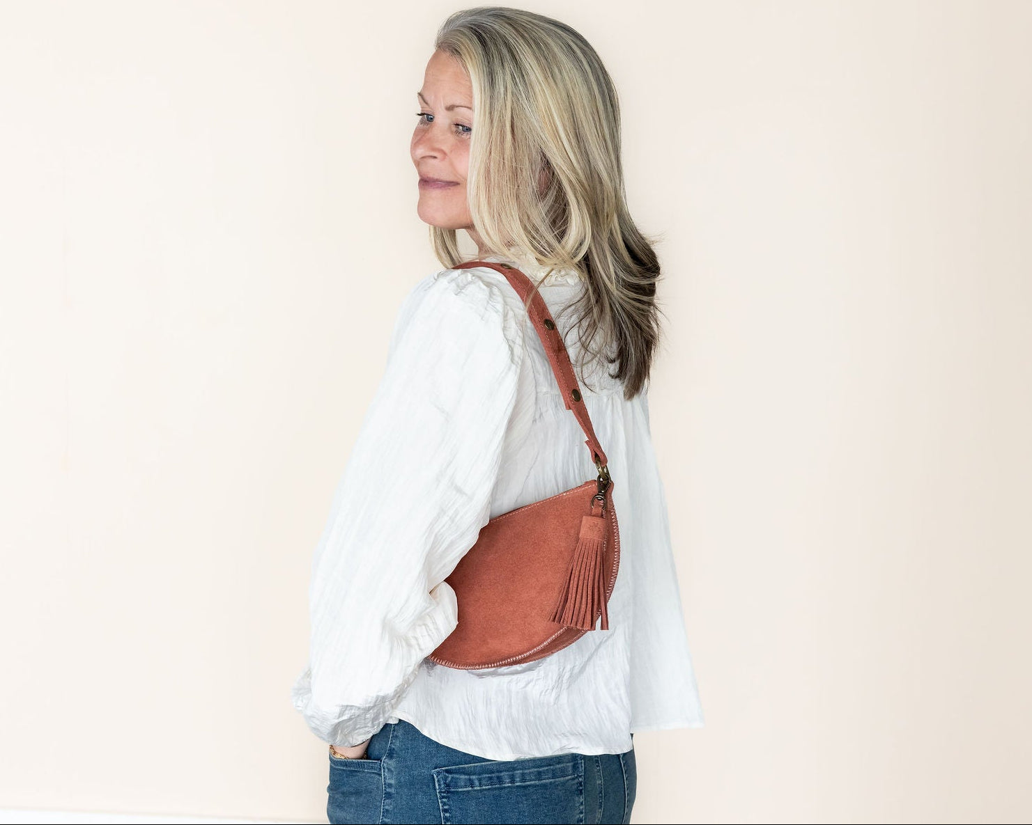 Woman wearing a white shirt and blue jeans with a mauve leather bag on a beige background
