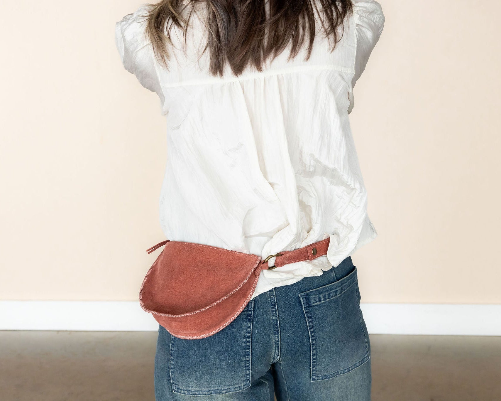 Person wearing a white blouse and blue jeans with a mauve leather fanny pack on a plain background