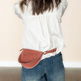 Person wearing a white blouse and blue jeans with a mauve leather fanny pack on a plain background