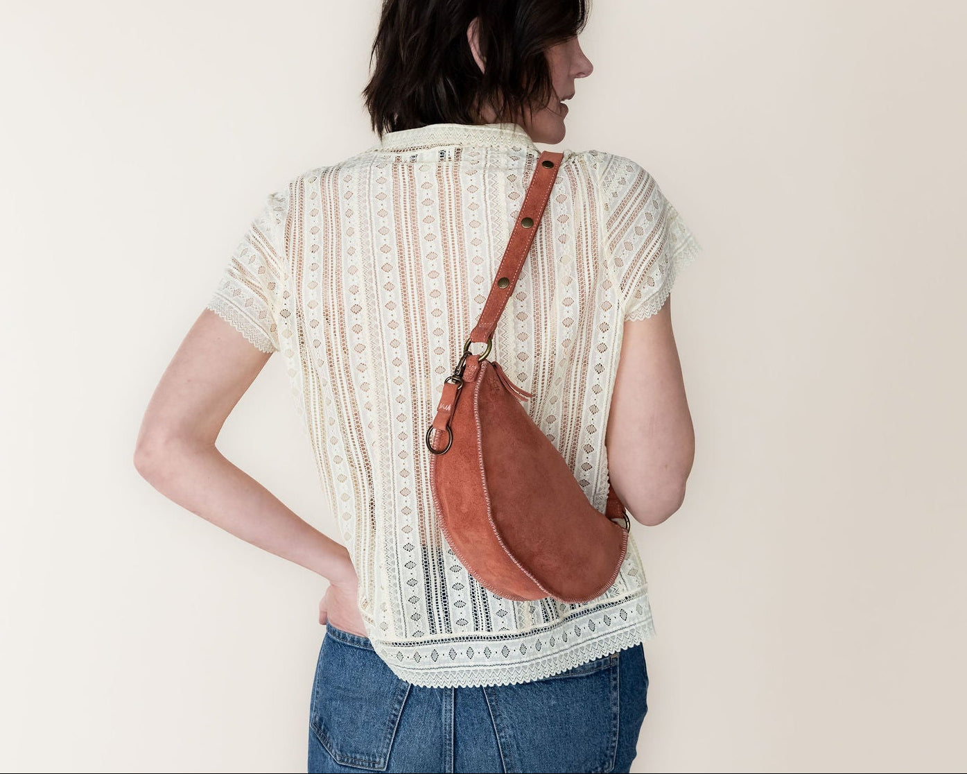 Person wearing a patterned shirt and jeans with a mauve leather bag over one shoulder against a plain background
