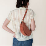 Person wearing a patterned shirt and jeans with a mauve leather bag over one shoulder against a plain background