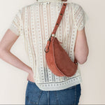 Person wearing a patterned shirt and jeans with a brown leather bag over one shoulder against a plain background