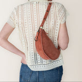 Person wearing a patterned shirt and jeans with a brown leather bag over one shoulder against a plain background