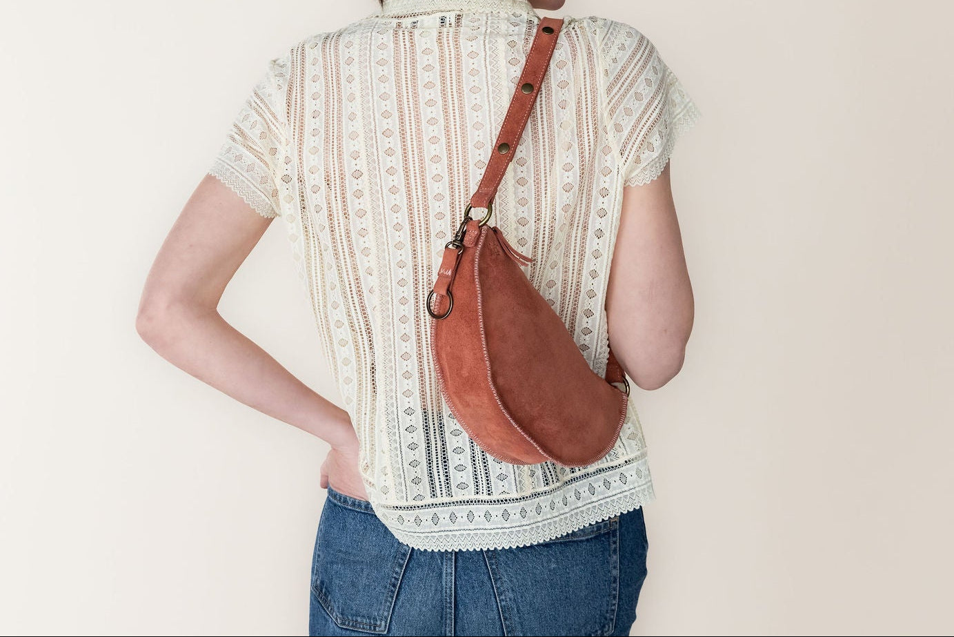 Person wearing a patterned shirt and jeans with a brown leather bag over one shoulder against a plain background