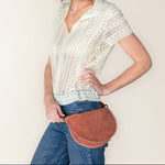 Woman wearing a white blouse and blue jeans, holding a brown leather bag against a plain background