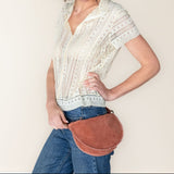 Woman wearing a white blouse and blue jeans, holding a brown leather bag against a plain background