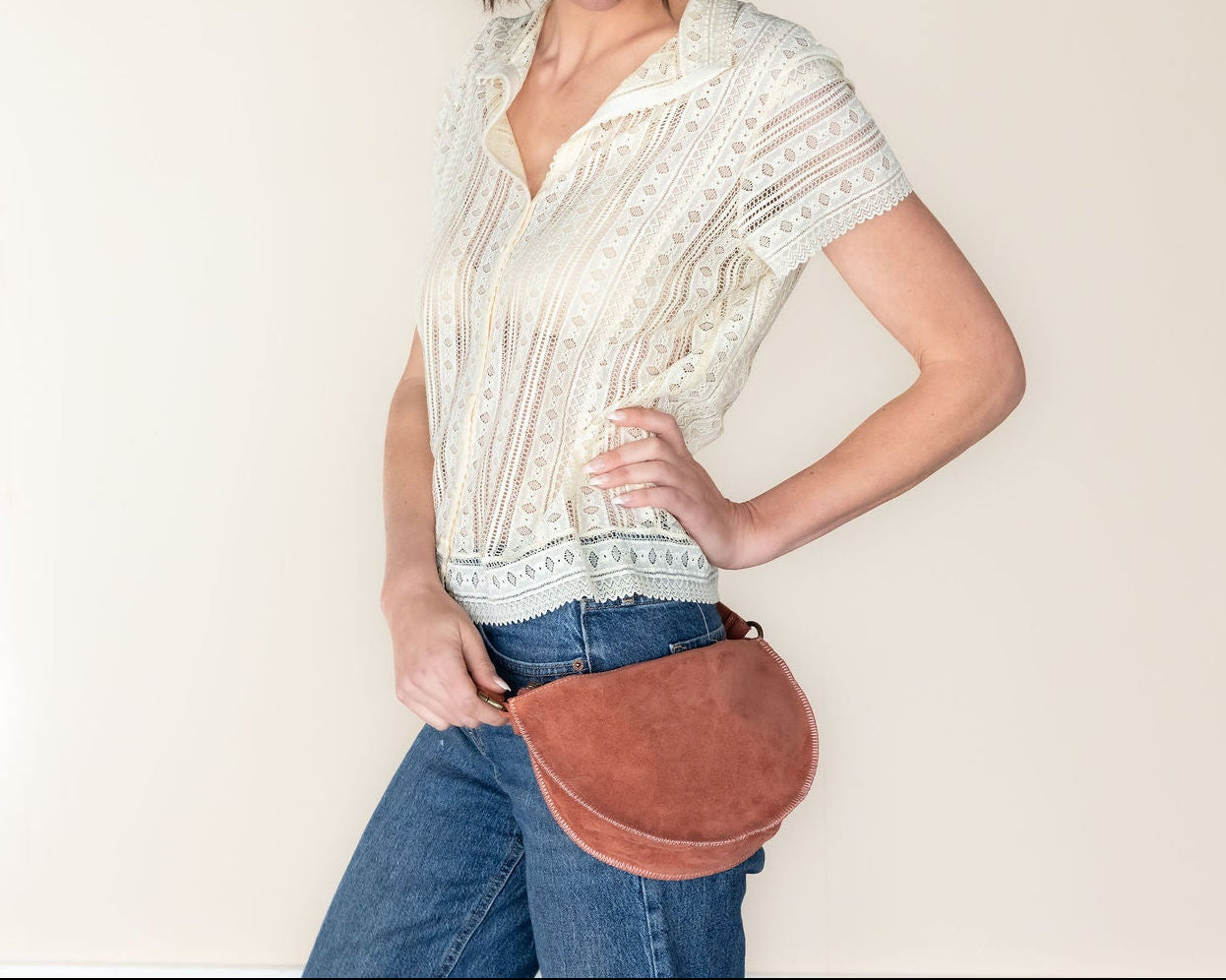 Woman wearing a white blouse and blue jeans, holding a brown leather bag against a plain background