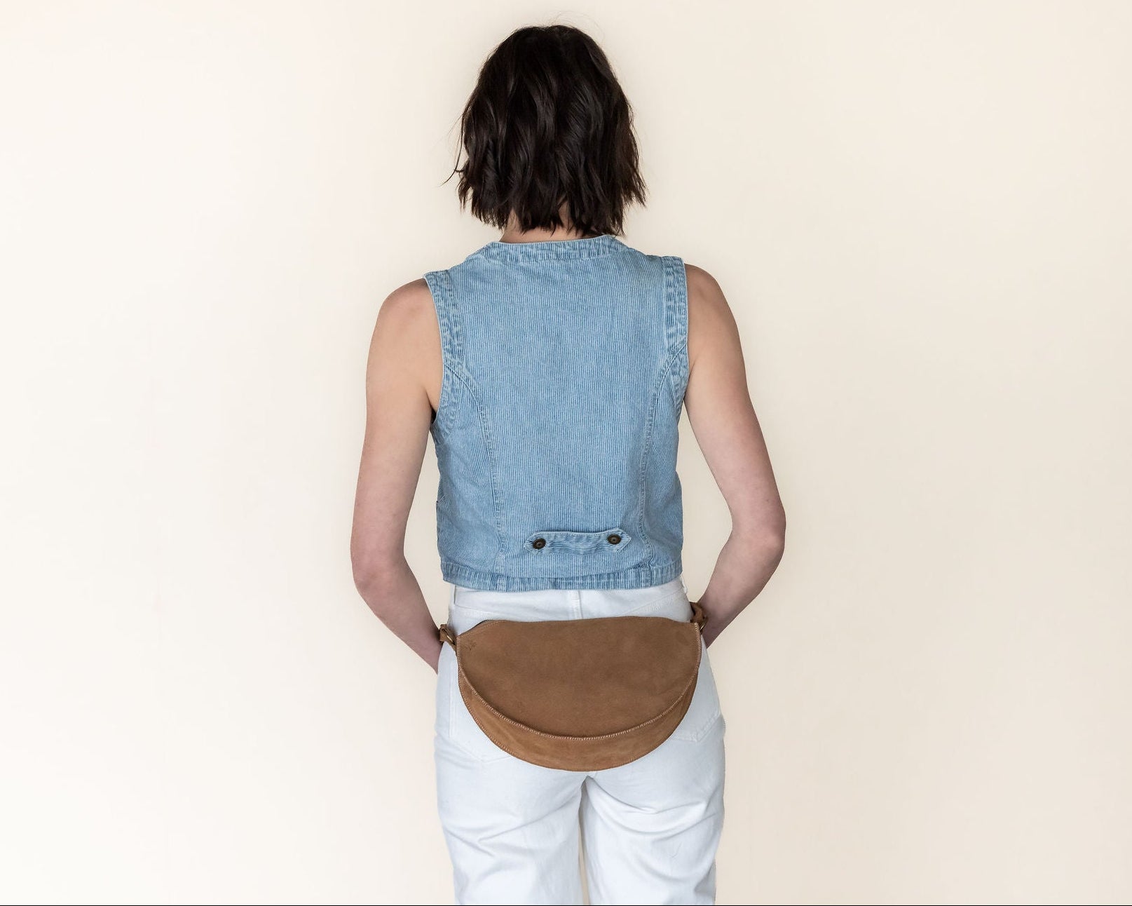 Person wearing a denim vest and white pants with a brown leather bag on a beige background