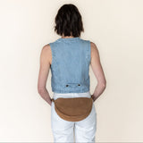 Person wearing a denim vest and white pants with a brown leather bag on a beige background