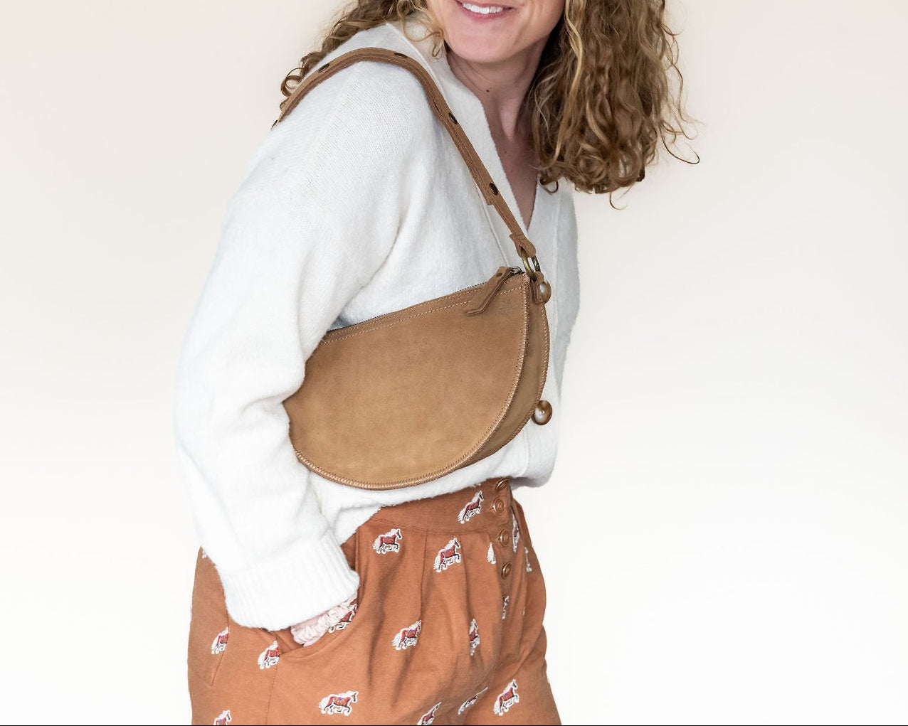 Person wearing a white shirt and brown patterned pants with a tan crossbody bag on a plain background