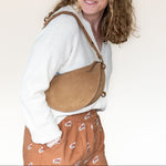 Person wearing a white shirt and brown patterned pants with a tan crossbody bag on a plain background