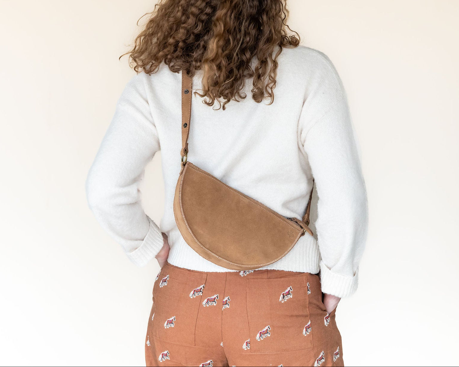 Person wearing a white sweater and patterned pants with a tan crossbody bag on a plain background
