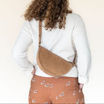 Person wearing a white sweater and patterned pants with a tan crossbody bag on a plain background
