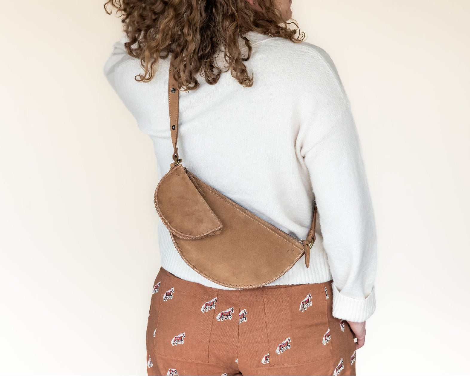 Person wearing a white sweater and brown patterned pants with a tan crossbody bag on a plain background