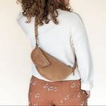 Person wearing a white sweater and brown patterned pants with a tan crossbody bag on a plain background