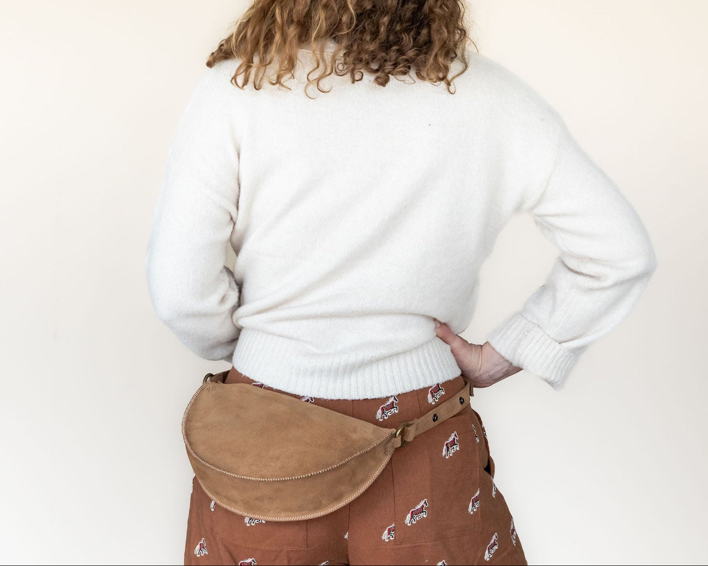 Person wearing a white sweater and brown skirt with a tan belt bag on a plain background