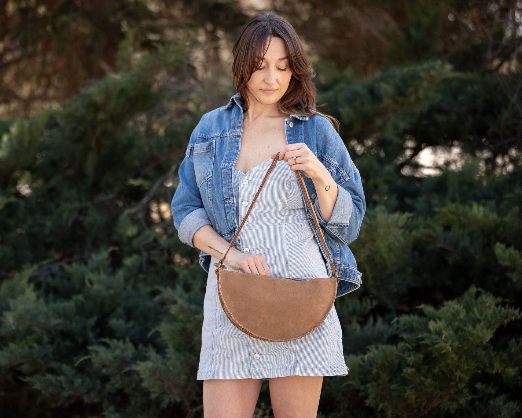 Woman holding a brown leather handbag outdoors with greenery in the background