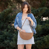 Woman holding a brown leather handbag outdoors with greenery in the background