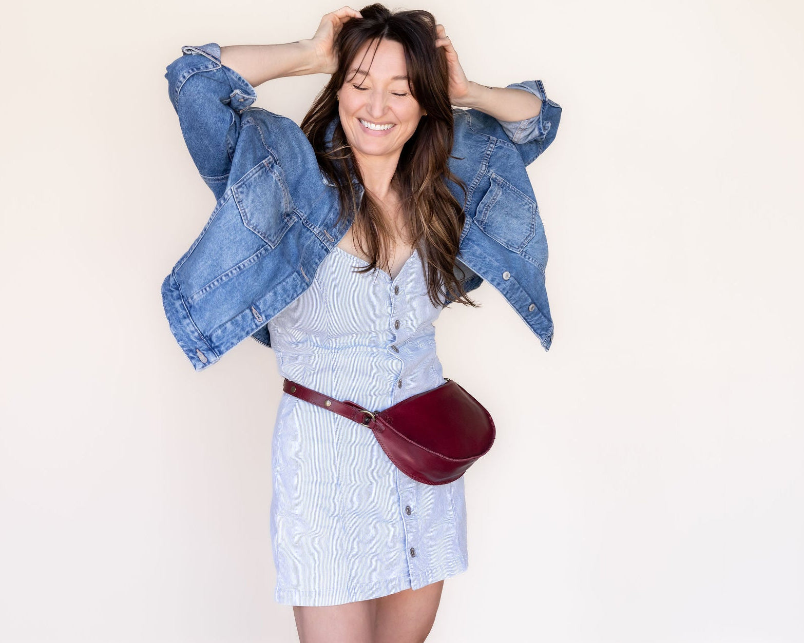 Woman wearing a denim jacket and white dress with a red bag against a plain background