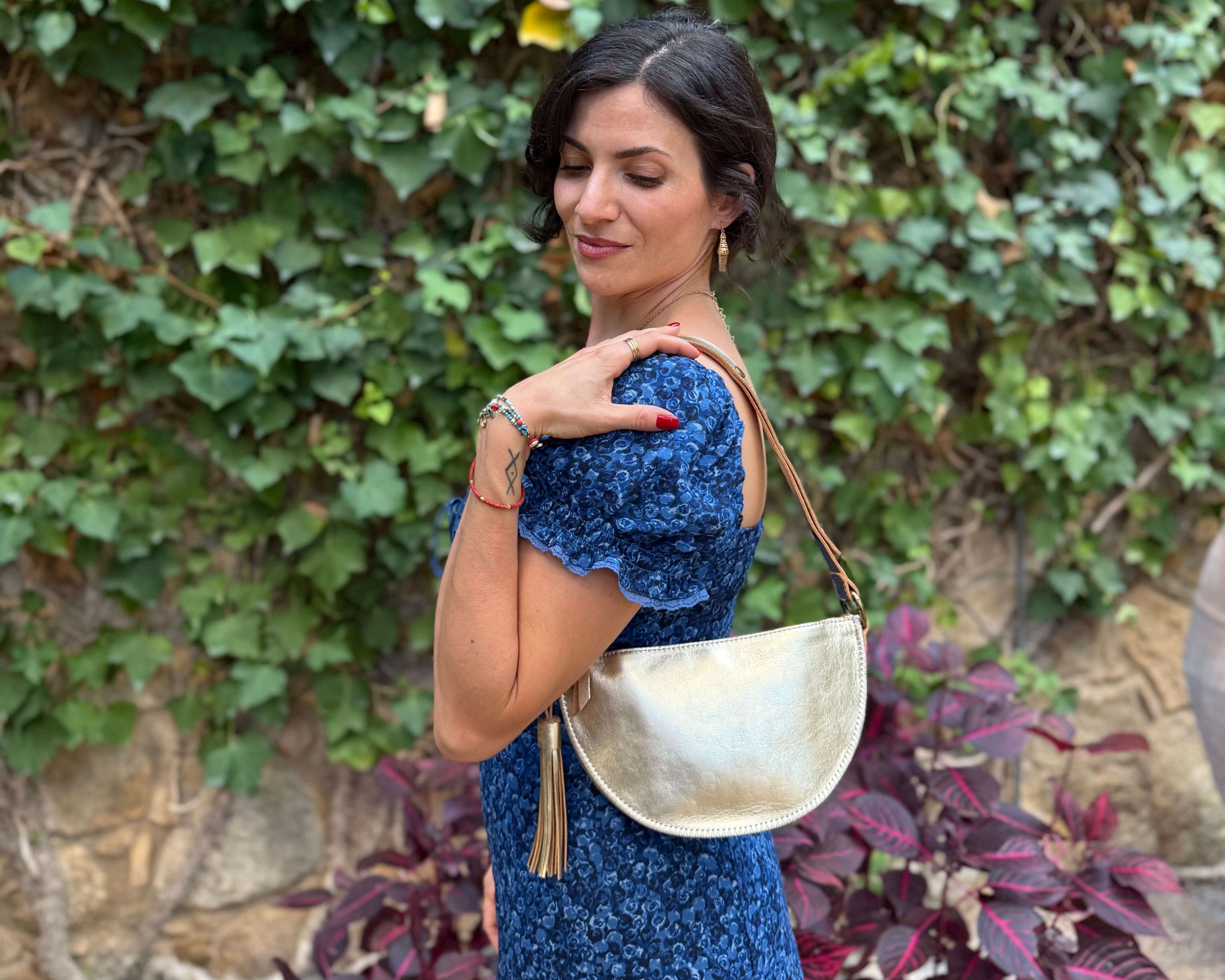 Woman in a blue dress holding a gold handbag against a green ivy wall.
