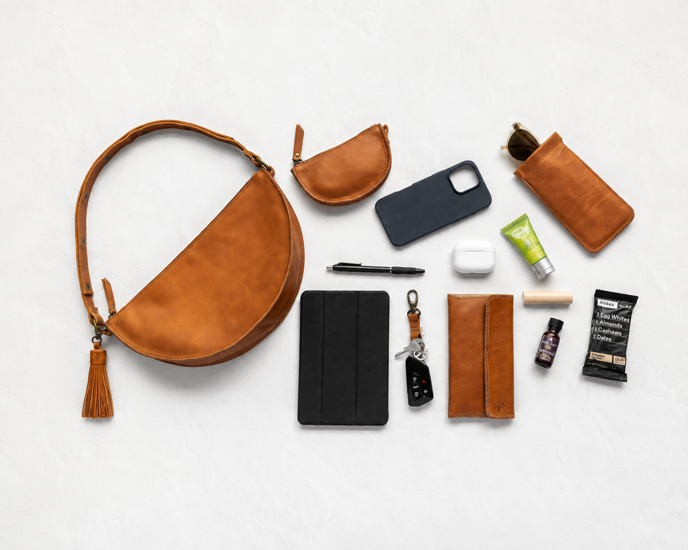 A curated flat-lay of brown leather accessories and small everyday items arranged on a light gray background, including a crescent shoulder bag with tassel, pouches, phone case, sunglasses holder, wallet, key fob, pen, earbuds case, and small toiletry items.